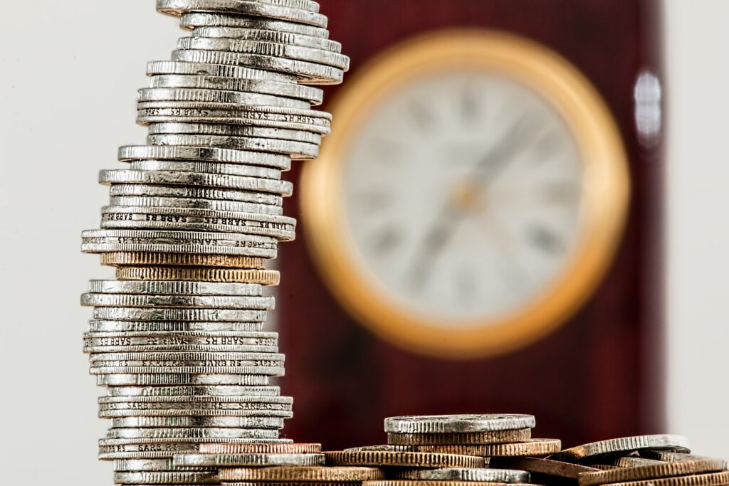 An image showing a pile of coins in the foreground and a clock out of focus in the background, depicting the connection between money and time.
